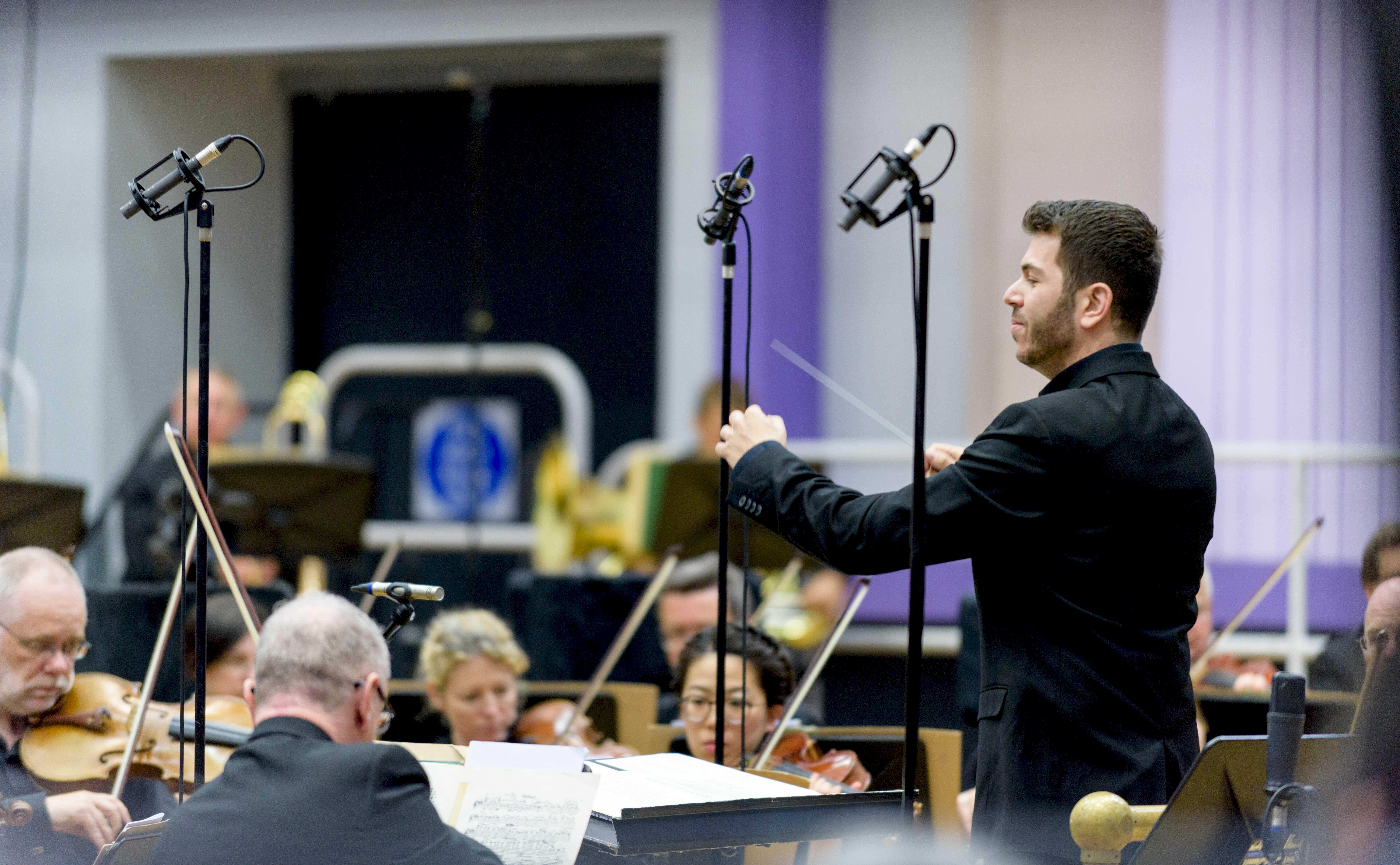 A conductor in a black shirt conducts the BBC Philharmonic Orchestra strings section.