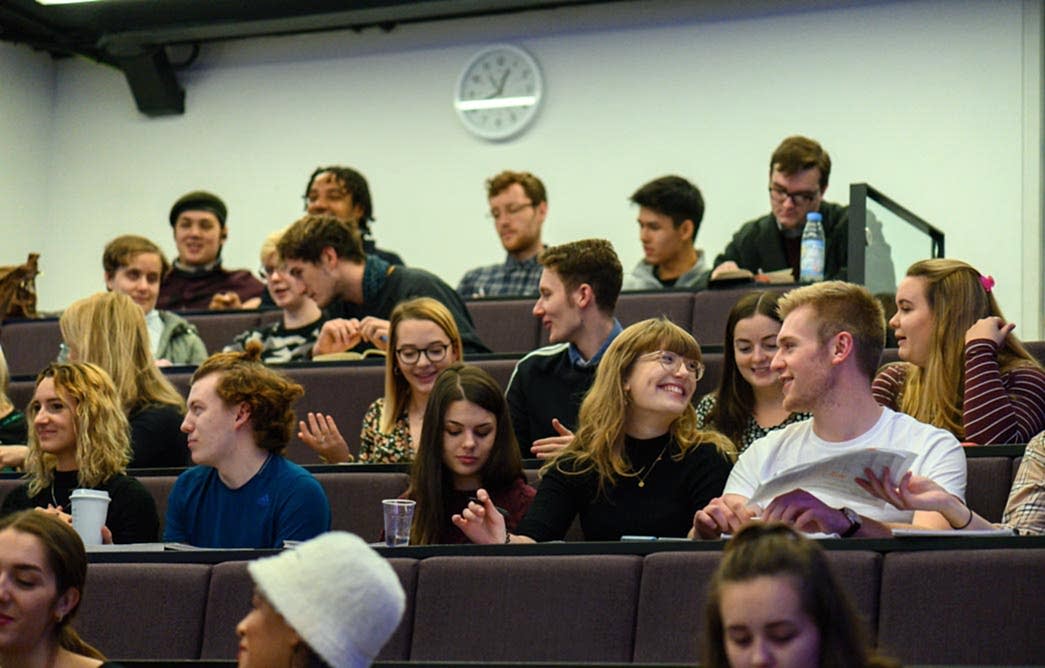 Students talk and laugh in groups as they sit in lecture theatre seating.