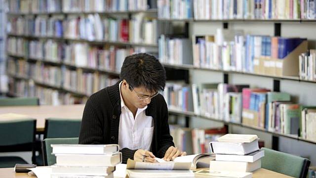 An RNCM student sits at a desk in a library reading and surrounded by books.
